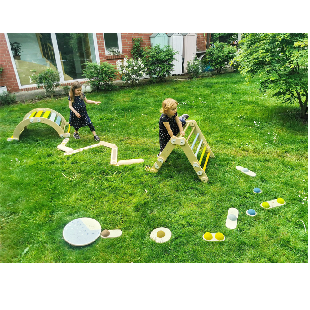 Zwei junge Mädchen spielen auf Balanciersteinen „Adventure“ und hölzernen Klettergerüsten in einem grasbewachsenen Hinterhof.