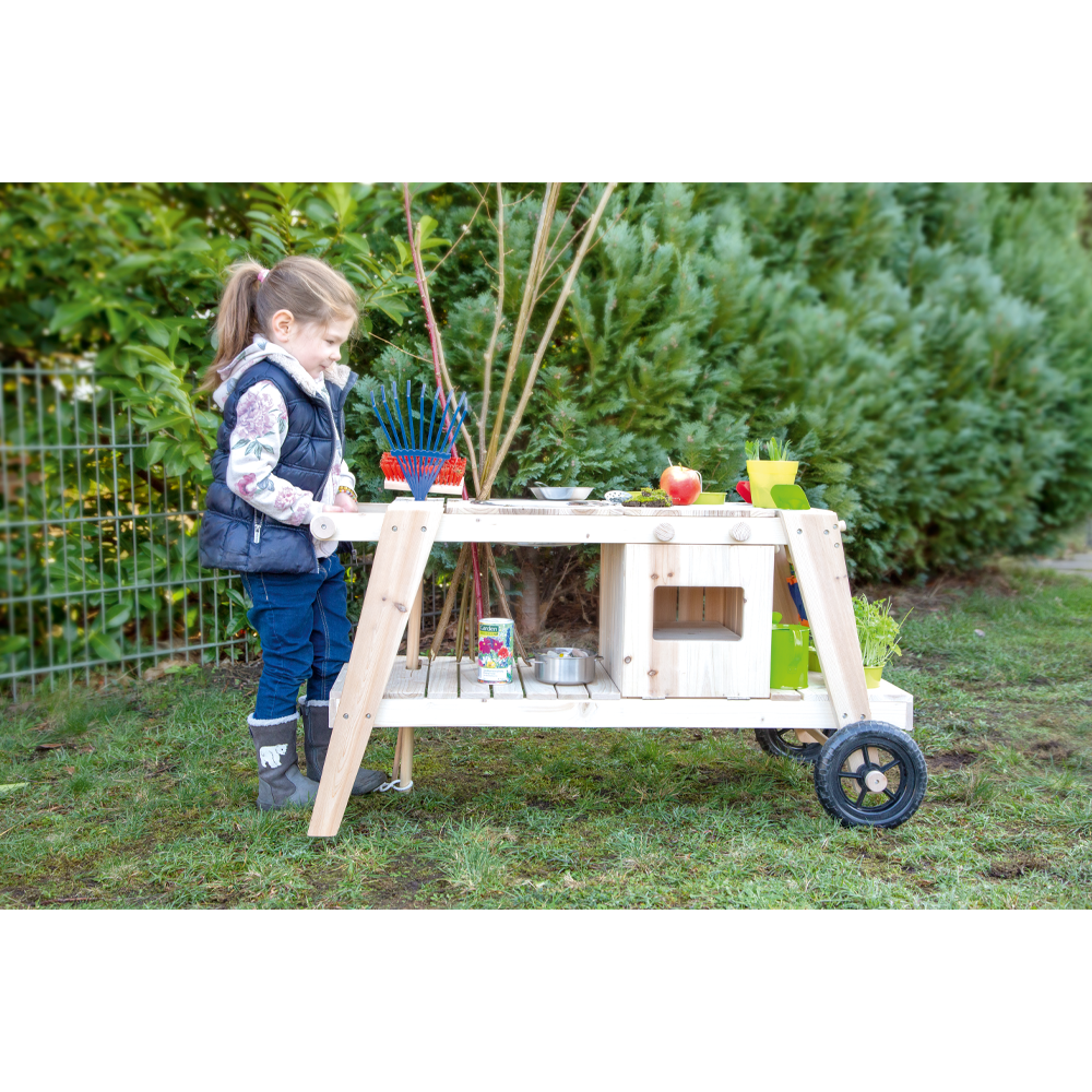 Ein junges Mädchen spielt mit der hölzernen Outdoor-Spielküche Matschküche in einem Garten mit grünen Büschen im Hintergrund.