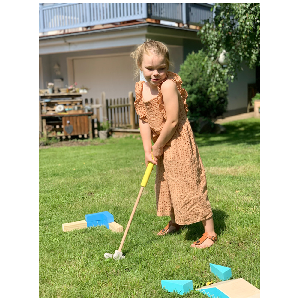 Ein junges Mädchen spielt an einem sonnigen Tag draußen Minigolf mit dem Minigolf-Set „Active“ und steht in einem braunen Kleid auf grünem Gras.