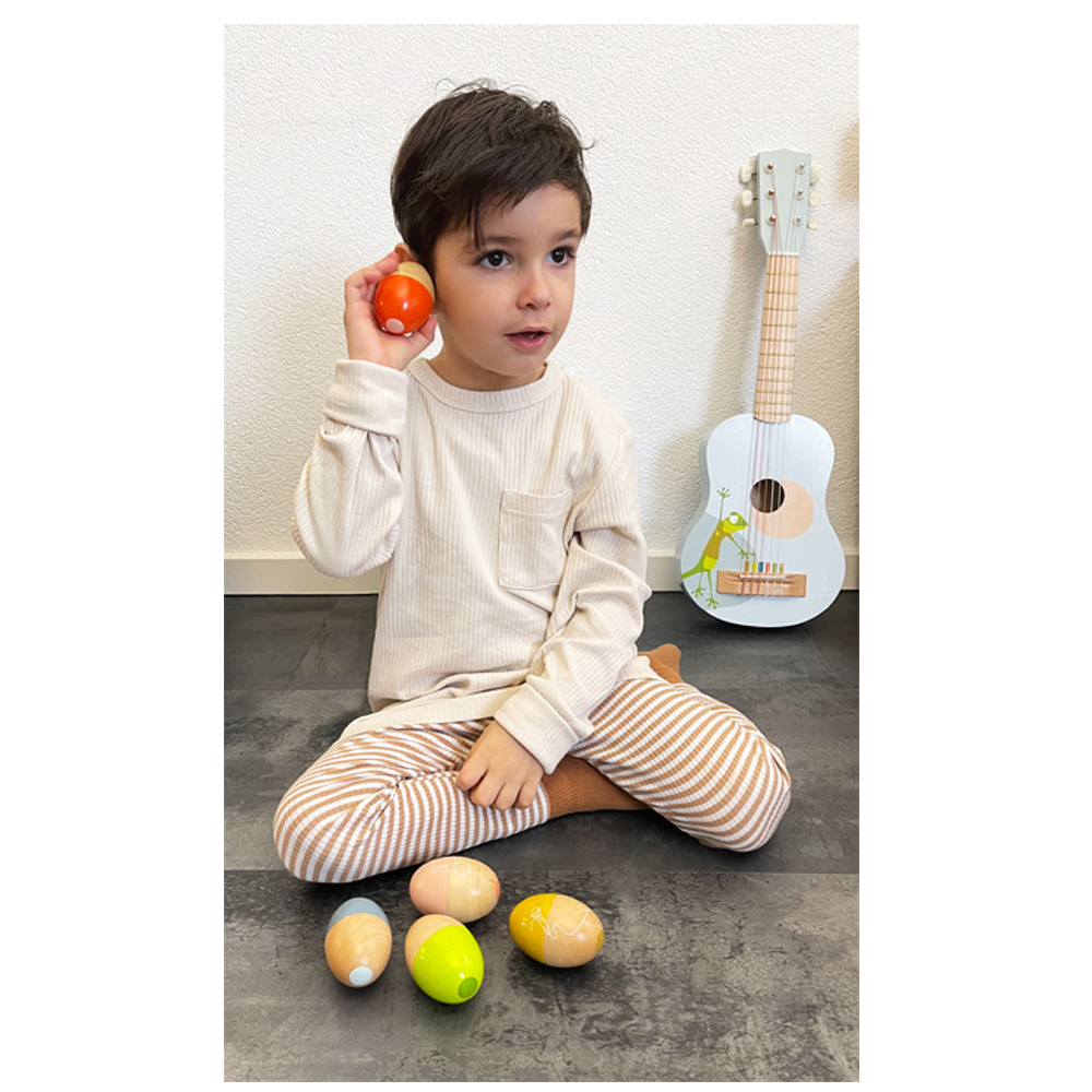 Ein kleiner Junge sitzt mit dem Rhythmusspielzeug „Groovy Beats“ von Musikeier auf dem Boden und hält einen eiförmigen Shaker an sein Ohr; hinter ihm steht eine kleine Gitarre.