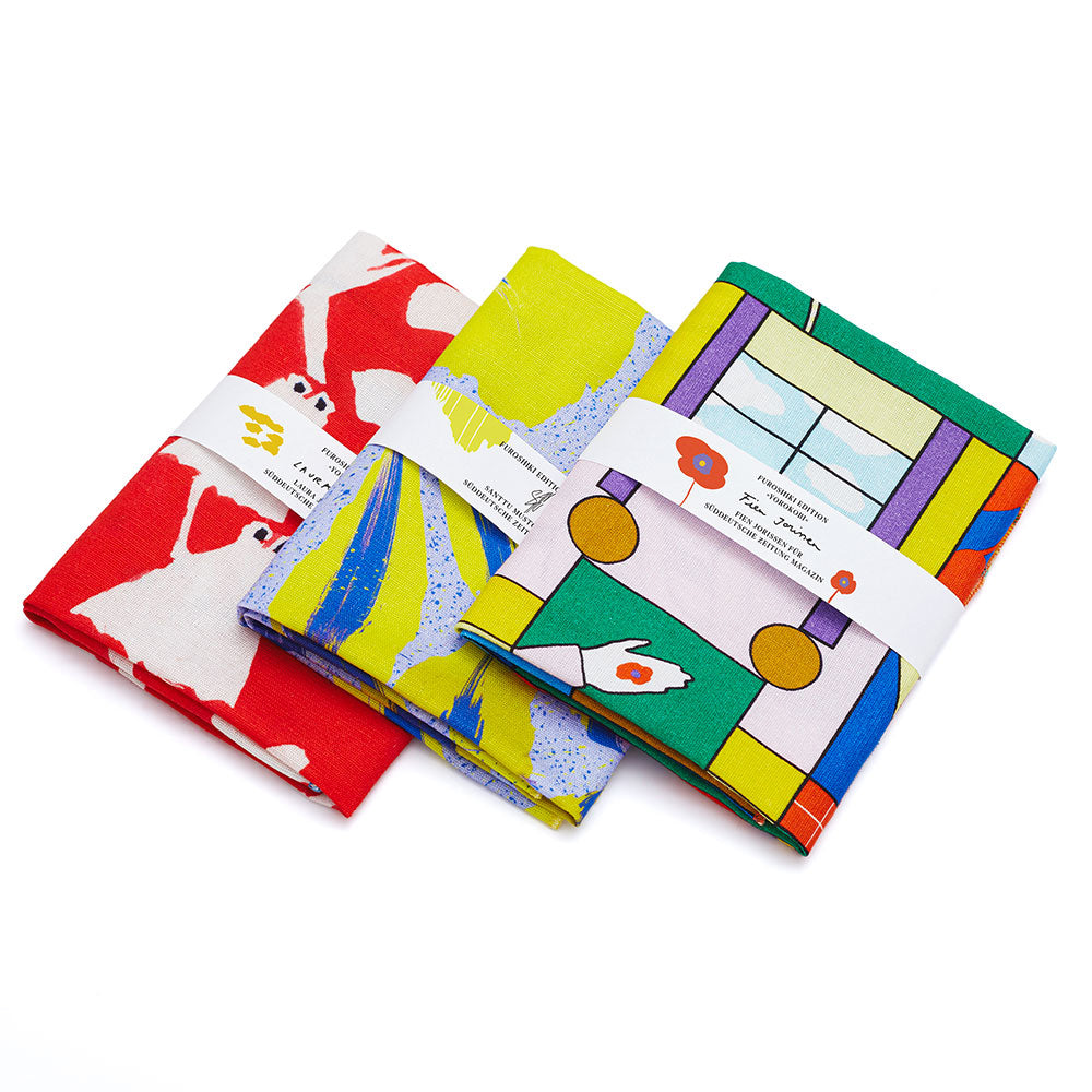 Drei bunte Falttücher mit abstrakten und geometrischen Mustern, jeweils mit einem weißen Papierband umwickelt.