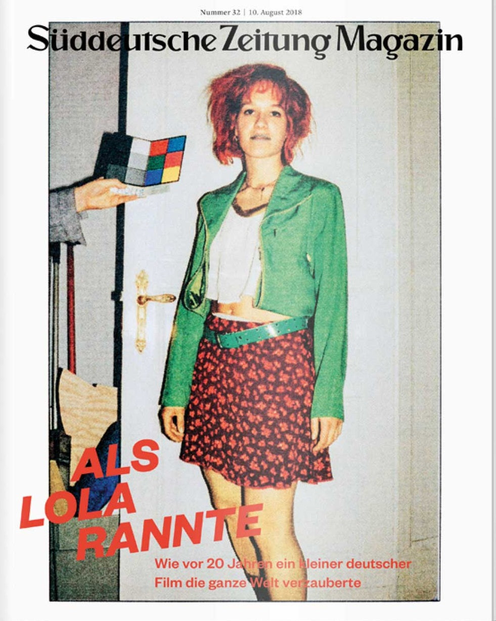 Eine Frau in grüner Jacke und rotem Rock steht an einer Wand; auf der Titelseite der Zeitschrift steht Als Lola rannte.