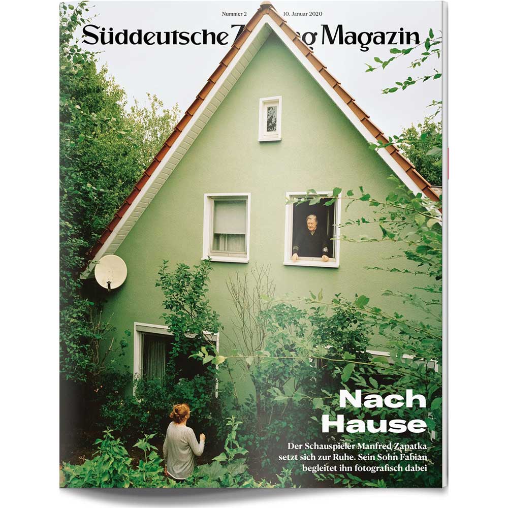 Ein grünes Haus mit zwei Personen: eine an einem Fenster im Obergeschoss und eine im Garten, umgeben von Grün.