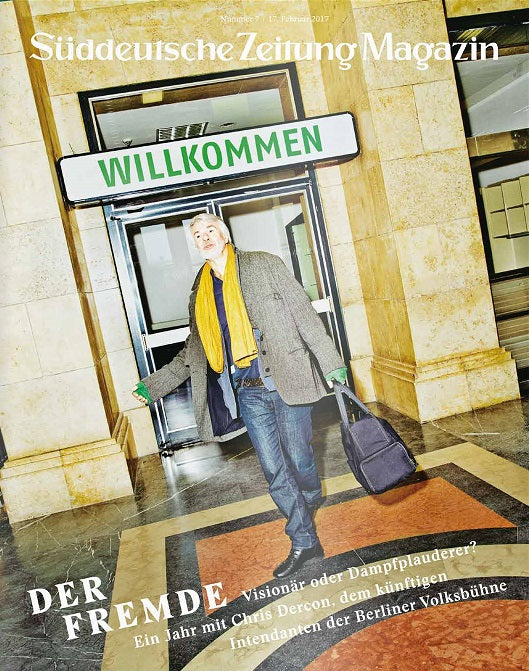 Ein Mann steht vor einem WILLKOMMEN-Schild mit einer Tasche und einem Schal in einer Halle.