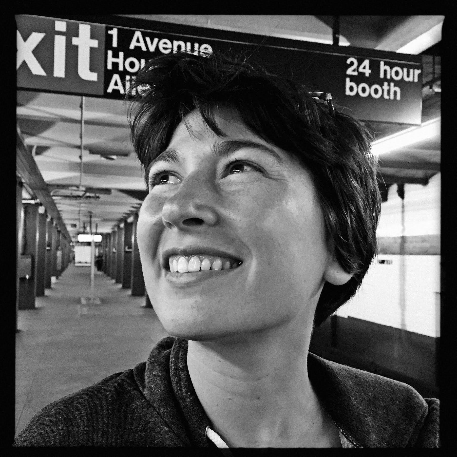 Eine Frau lächelt in einer U-Bahn-Station unter einem Schild mit der Aufschrift „Ausgang“, „1 Avenue“ und „24-Stunden-Schalter“.