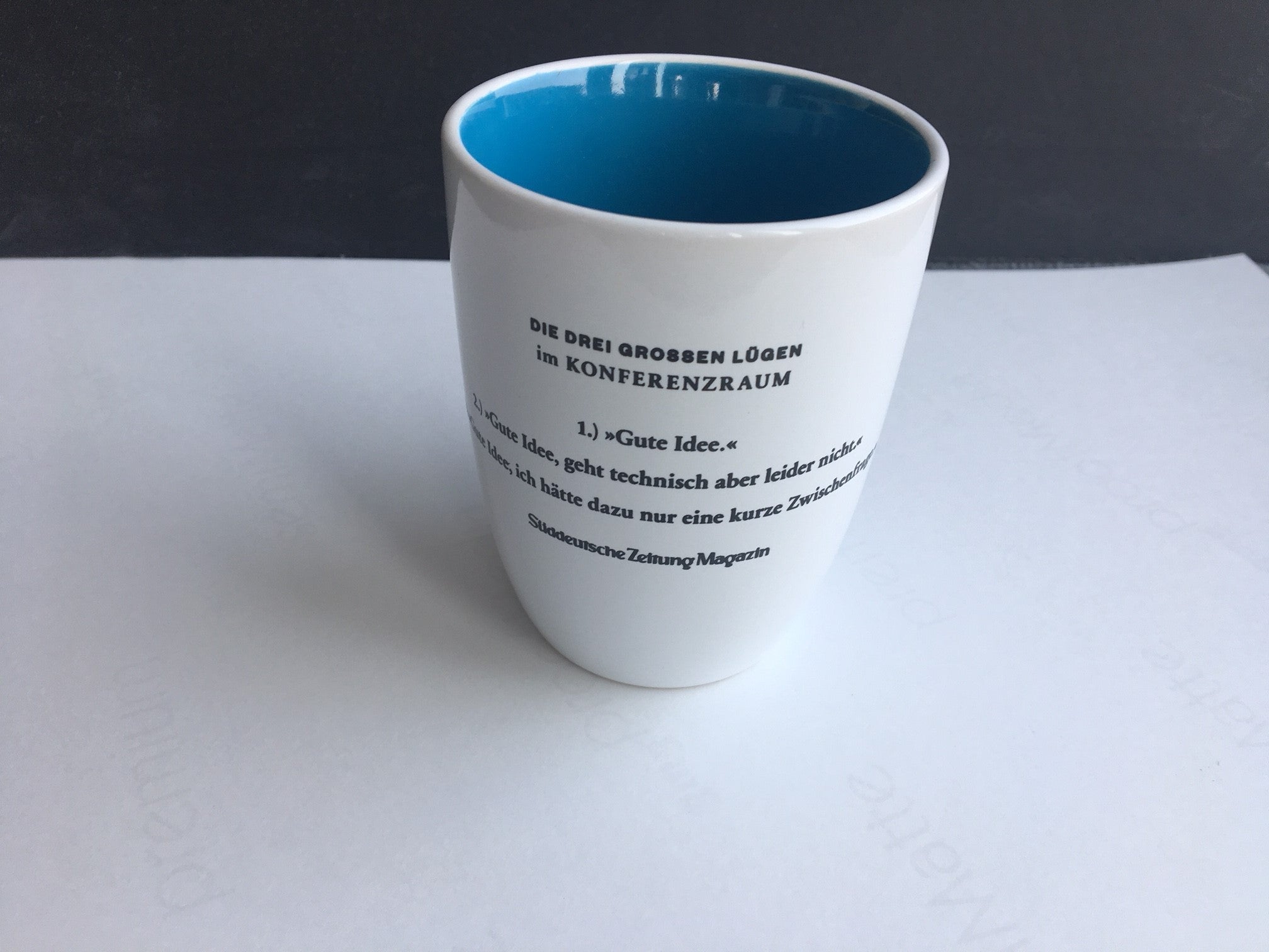 Eine weiße Tasse mit deutschem Aufdruck, innen blau, auf einem weißen Untergrund platziert.