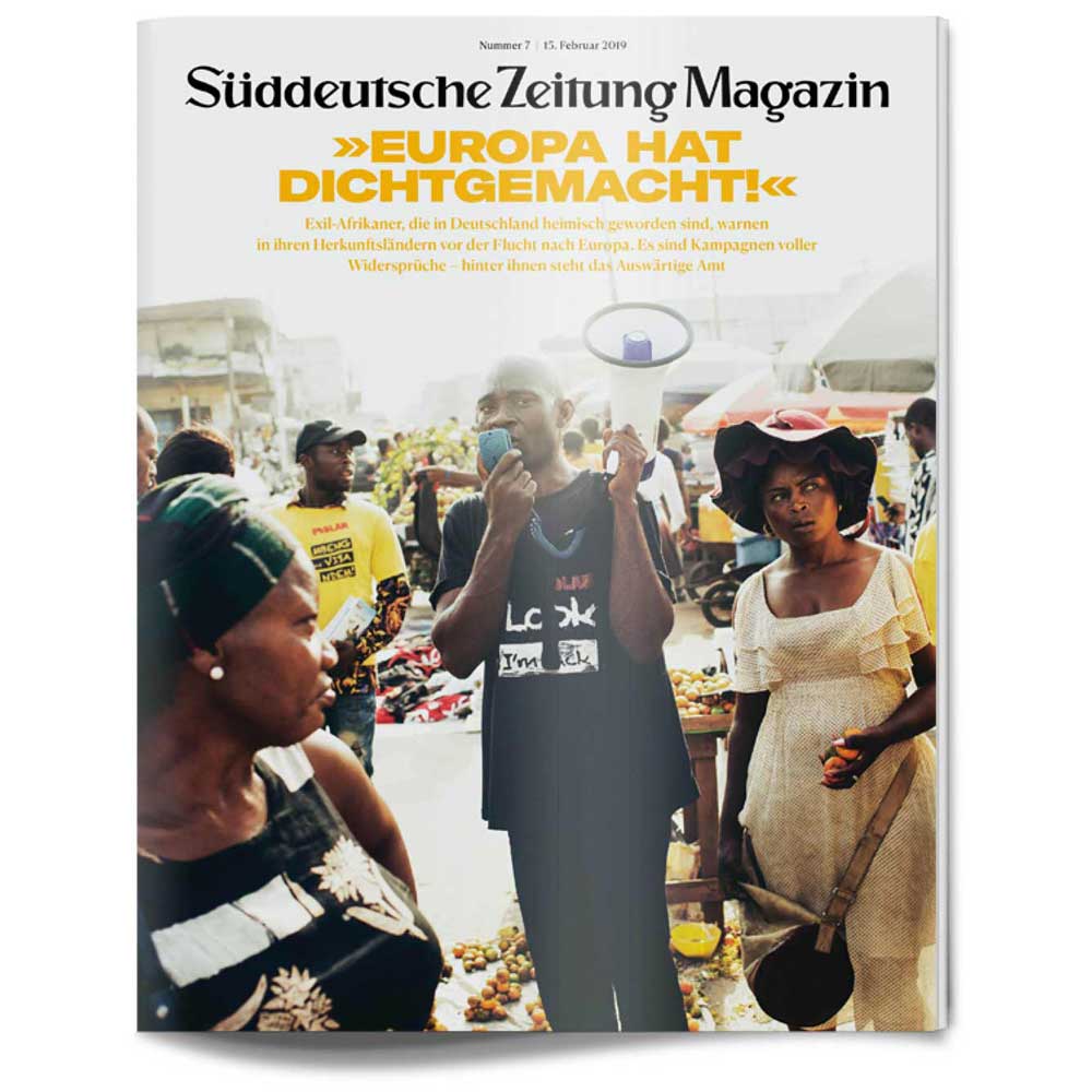 Menschen in farbenfroher Kleidung stehen auf einem lebhaften Markt im Freien und tauschen sich aus, wie auf dem Titelbild einer Zeitschrift zu sehen ist.