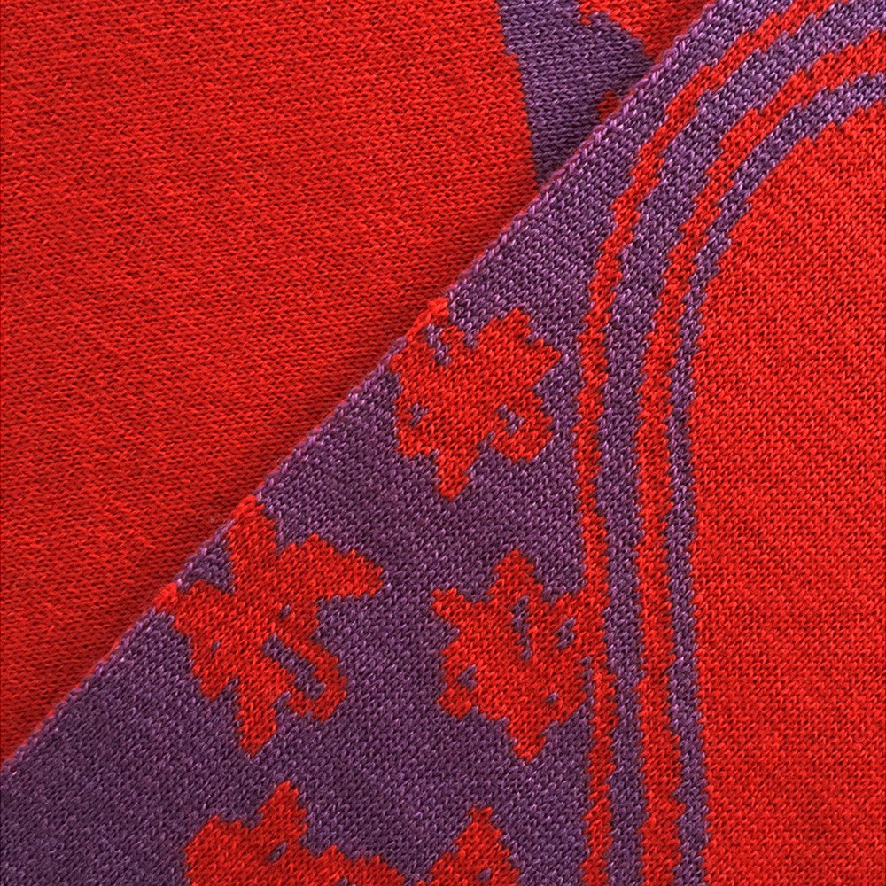 Nahaufnahme eines roten und violetten Strickstoffs mit diagonal verlaufenden floralen und geschwungenen Mustern.