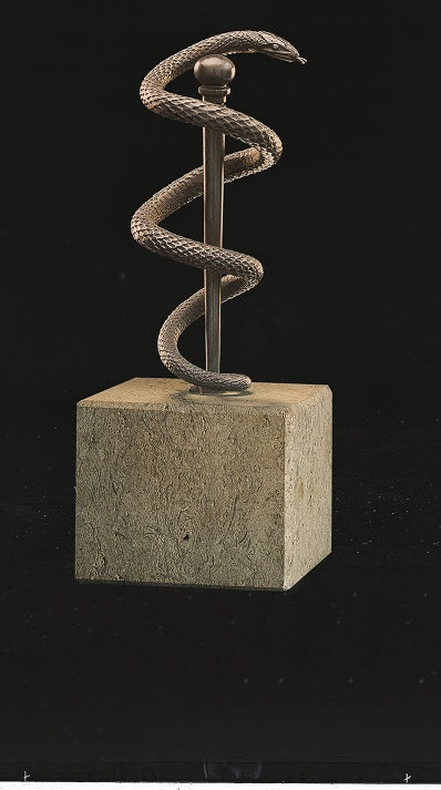 Auf einem quadratischen Steinsockel windet sich vor schwarzem Hintergrund eine Metallschlange um einen vertikalen Stab.