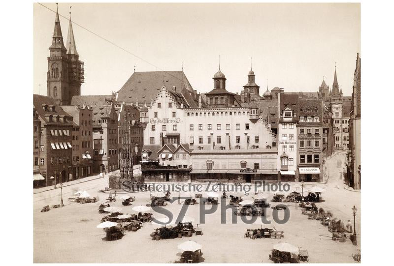 Historischer Stadtplatz mit Tischen und Stühlen im Freien, umgeben von alten Gebäuden und Kirchtürmen im Hintergrund.