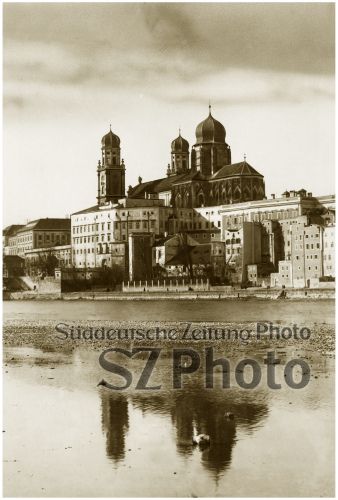 Historisches Stadtbild mit Kuppelkirche, großen Gebäuden und Flussreflexion unter bewölktem Himmel.