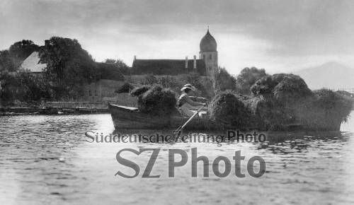 Eine Person rudert ein kleines, mit Heu beladenes Boot auf einem See, mit Gebäuden und einer Kirche im Hintergrund.