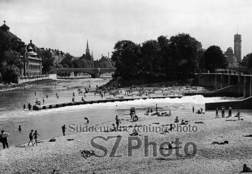 Menschen schwimmen und entspannen an einem Fluss in einer Stadt, mit historischen Gebäuden und Bäumen im Hintergrund.