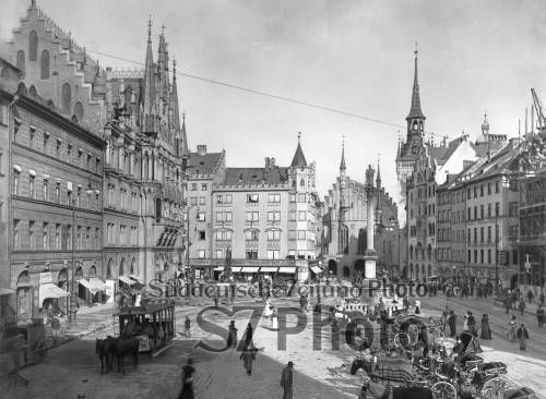 Historischer Stadtplatz mit Pferdekutschen, Fußgängern und Gebäuden im gotischen Stil an einem sonnigen Tag.