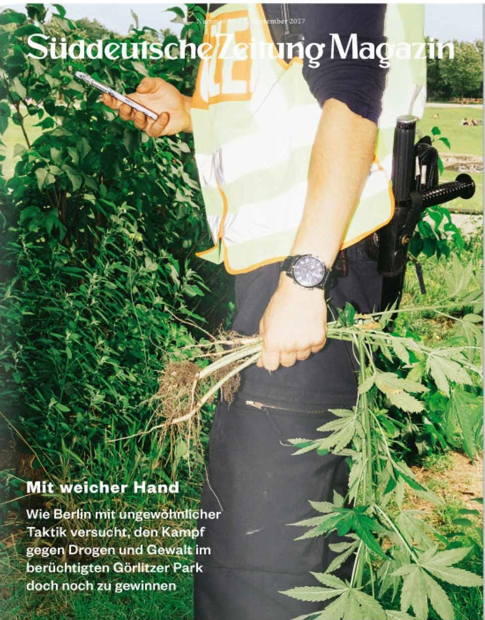 Eine Person in einer Sicherheitsweste hält ein Telefon und entwurzelte Cannabispflanzen in der Hand; eine Pistole im Halfter ist sichtbar.