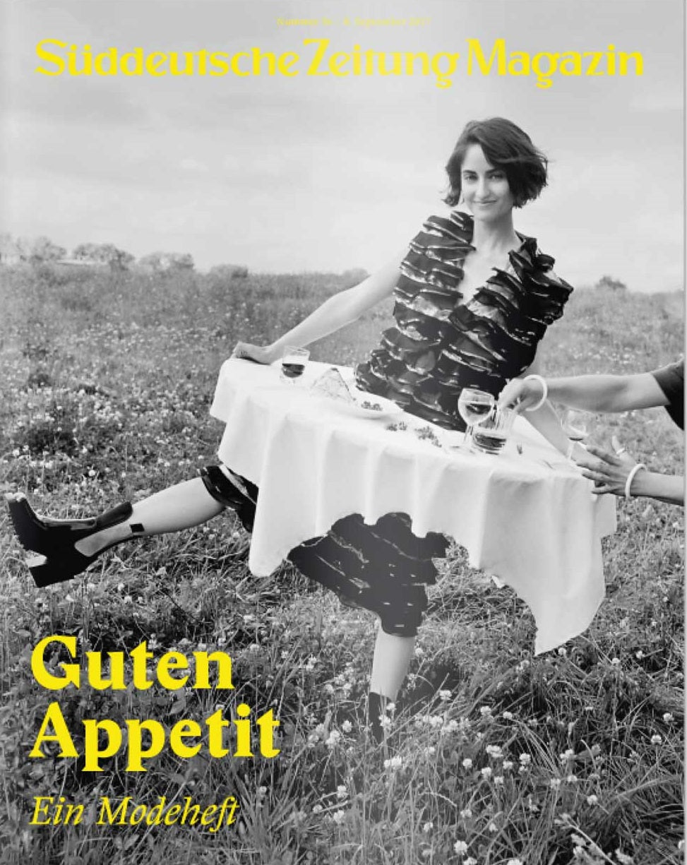 Eine Frau in einem Kleid hält einen mit Geschirr gedeckten Tisch in einem Feld; in gelber Schrift steht Guten Appetit.