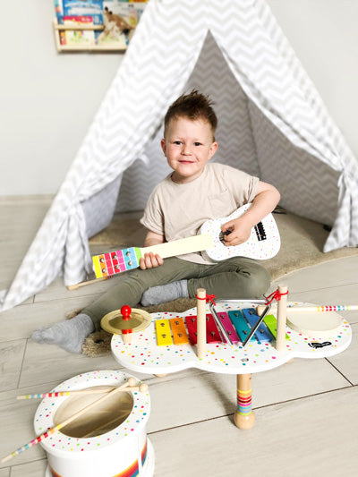 Ein kleines Kind sitzt vor einem Spielzelt und hält die Spielzeuggitarre „Gitarre Sound“. In der Nähe stehen andere Spielzeugmusikinstrumente.