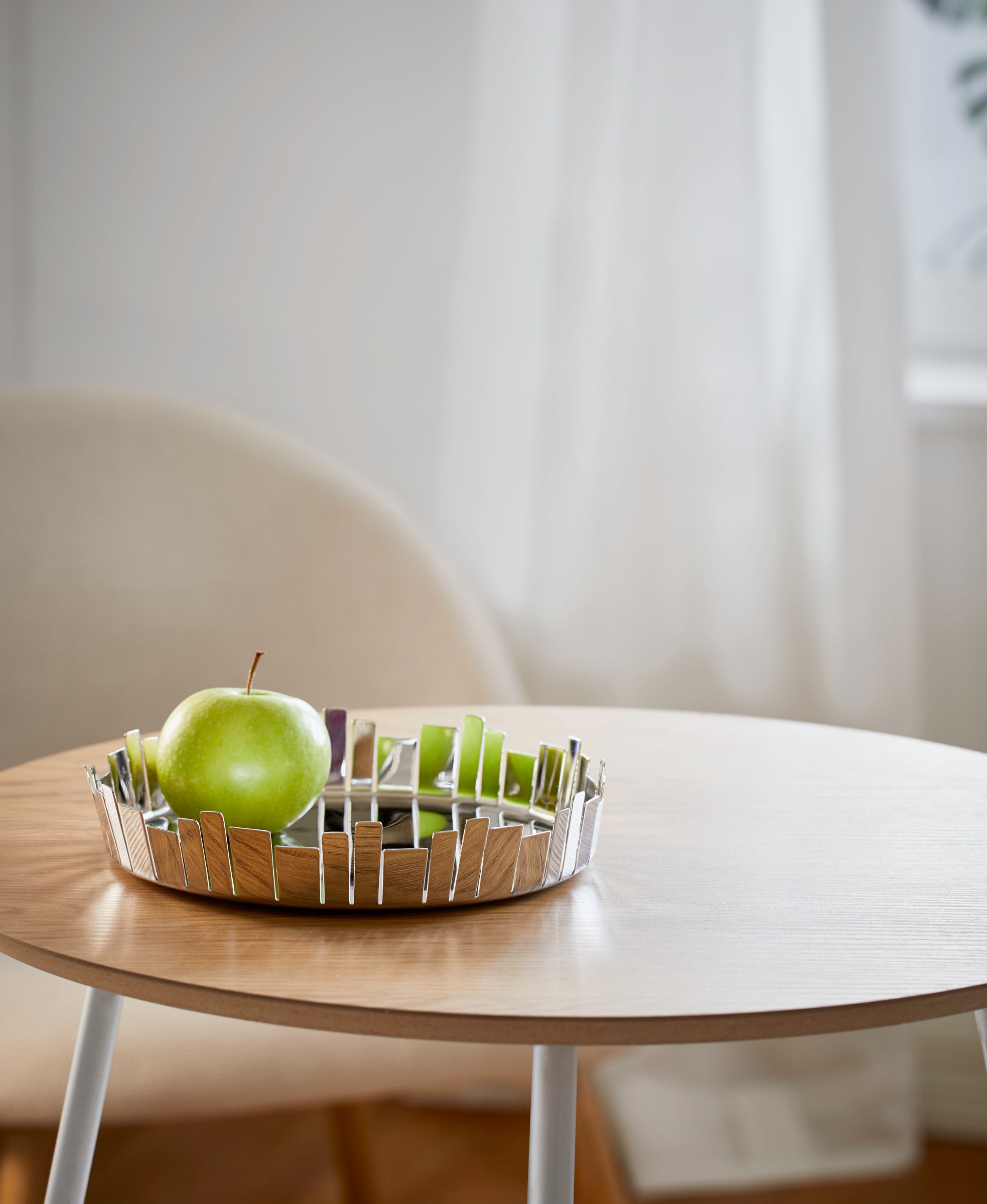 Ein grüner Apfel und Apfelscheiben in einer Metallschale auf einem Holztisch, im Hintergrund ein Stuhl.
