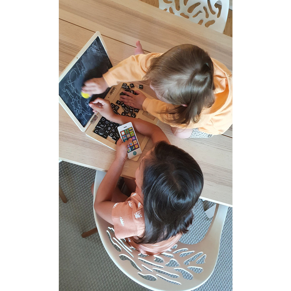 Zwei Kinder sitzen an einem Tisch, eines malt auf dem Holz-Laptop mit Magnet-Tafel, das andere spielt mit Buchstabensteinen und einem Spielzeugtelefon.