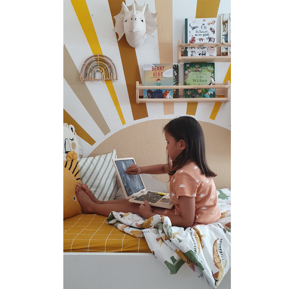 Ein junges Mädchen sitzt mit dem Holz-Laptop mit Magnet-Tafel auf einem Bett, im Hintergrund sind Bücher und Wanddekor zu sehen.