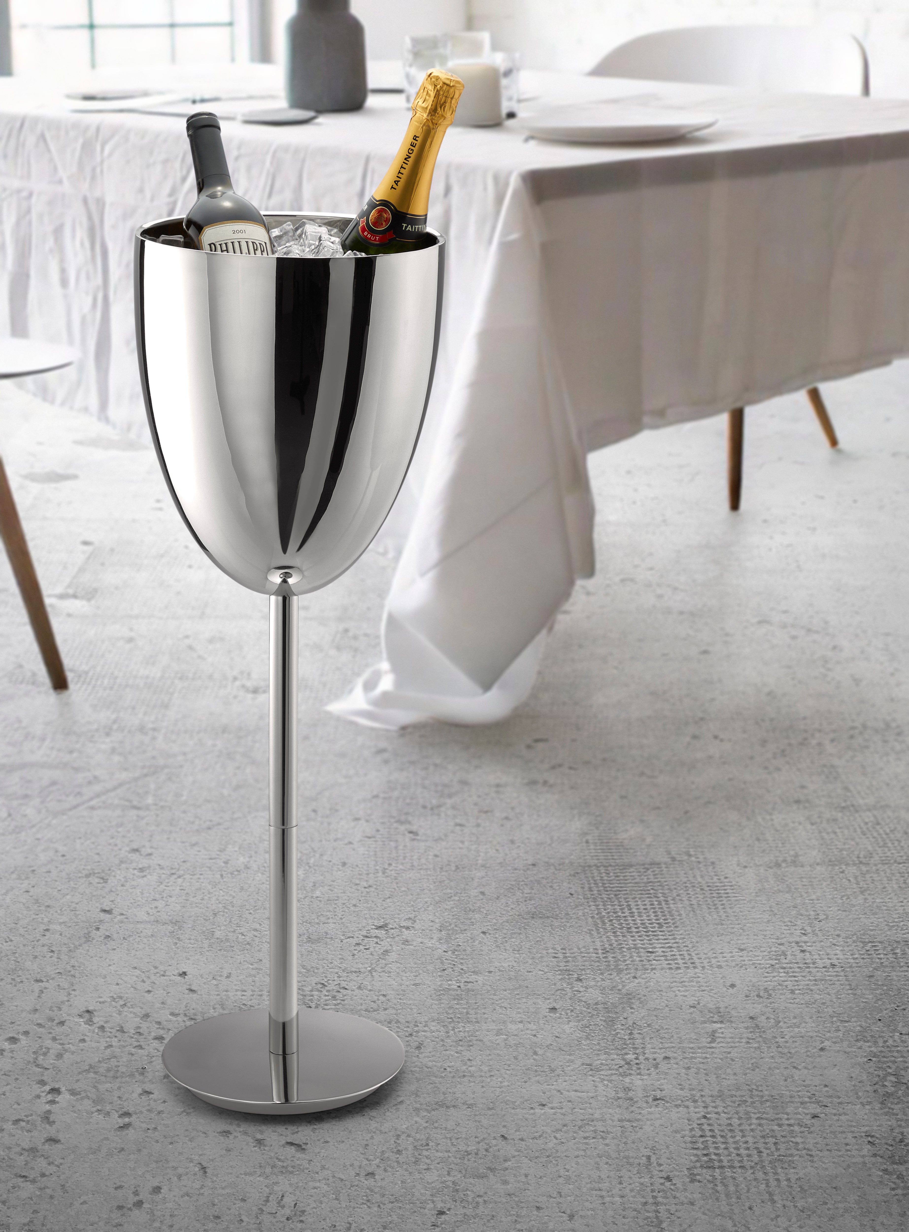 Neben einem Esstisch steht ein großer, silberner Weinkühler mit Eis, in dem sich eine Weinflasche und eine Champagnerflasche befinden.