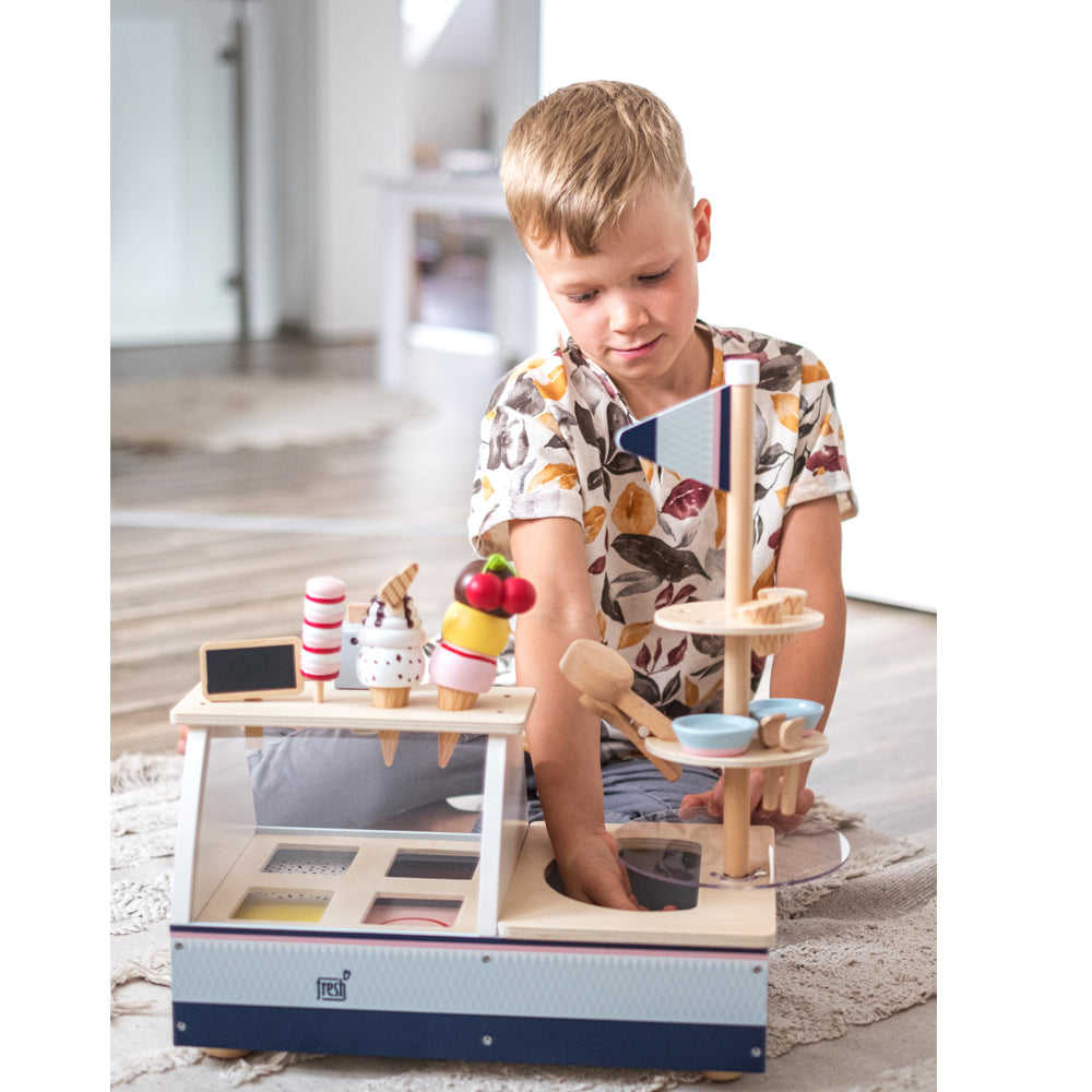 Kleiner Junge spielt mit dem Eisstand „Fresh“ aus Holz auf dem Boden eines hellen Raumes.