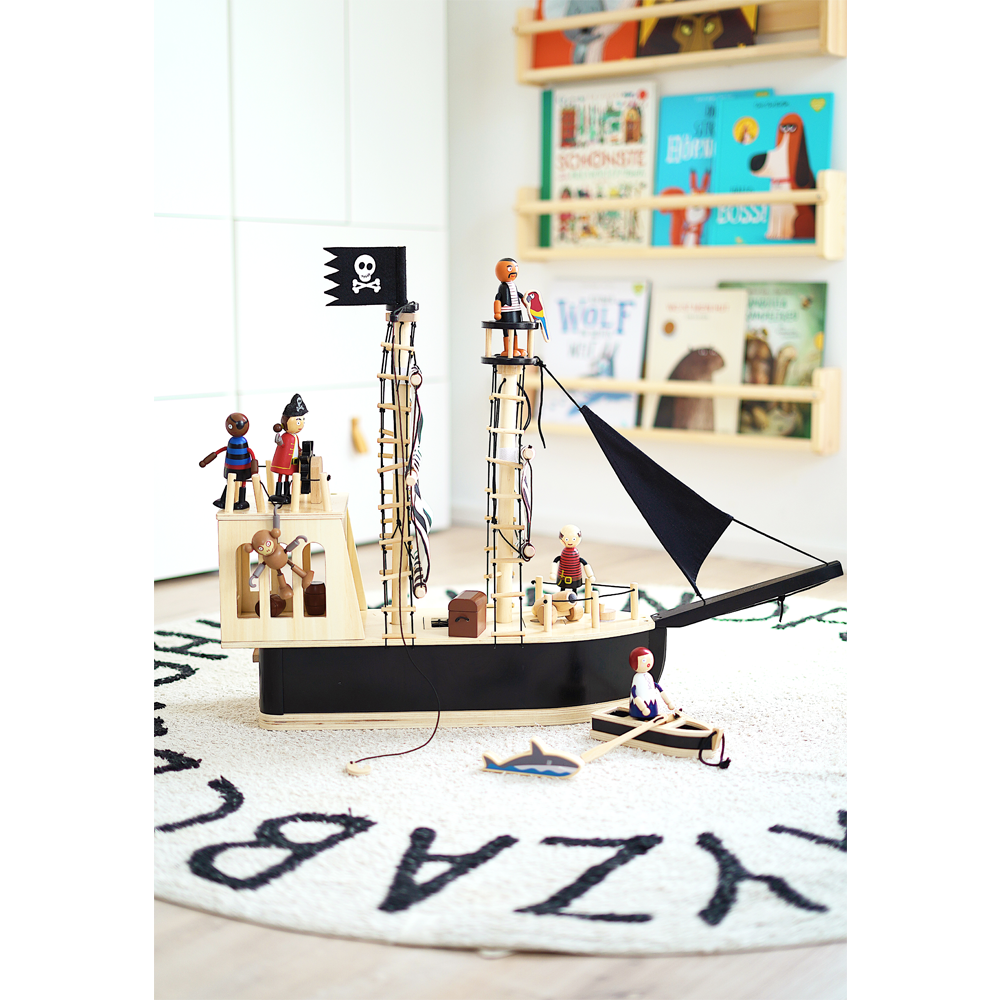 Das hölzerne Piratenschiff-Spielzeug mit Figuren steht auf einem runden Teppich, im Hintergrund sind Kinderbücherregale zu sehen.