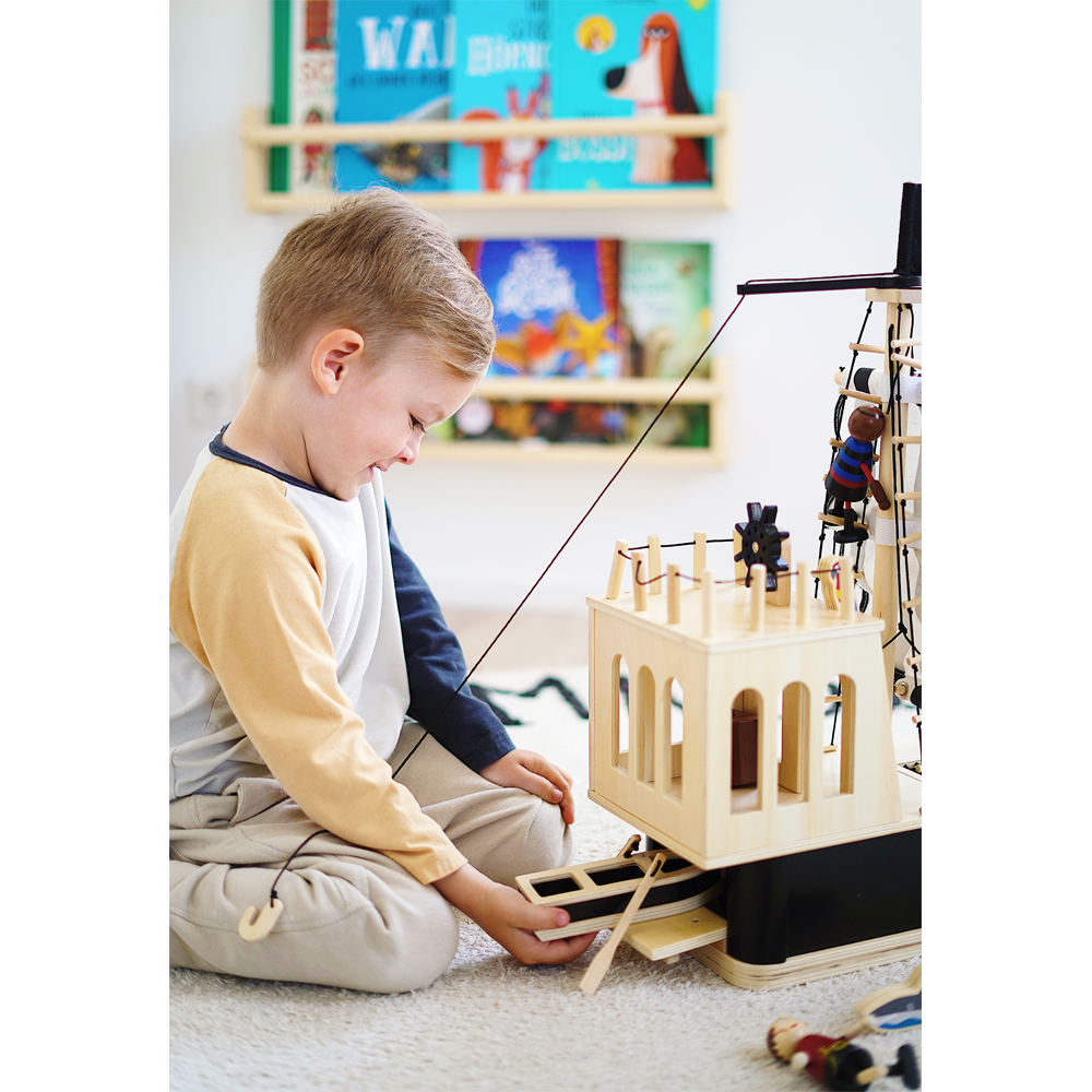 Ein kleiner Junge spielt mit dem hölzernen Piratenschiff-Spielzeug, im Hintergrund sind bunte Bücher ordentlich auf Regalen angeordnet.