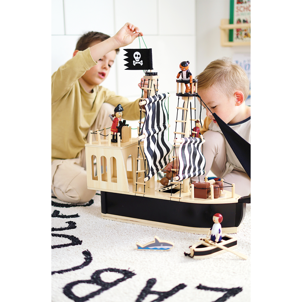 Zwei Kinder spielen mit dem Piratenschiff auf einem weißen Teppich, umgeben von Piratenfiguren und einem kleinen Boot.