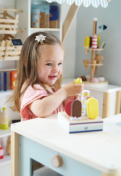 Lächelndes junges Mädchen spielt in einer Spielküche mit den bunten „frischen“ Spielzeugdesserts von Eis am Stiel.