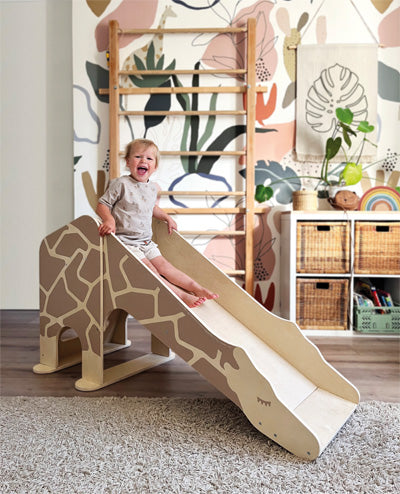 Ein lächelndes Kleinkind rutscht in einem farbenfroh dekorierten Spielzimmer die Indoor Rutsche Giraffe „Wildlife“ hinunter.