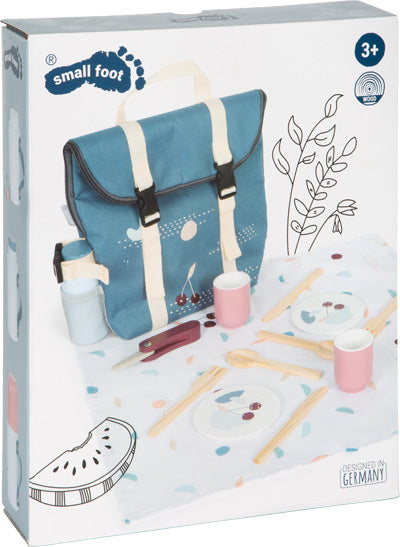 Box mit Picknickrucksack „Lecker“ mit einer blauen Tasche, Bechern, Besteck, Tellern und Spiellebensmitteln, die auf einem Tuch angeordnet sind.