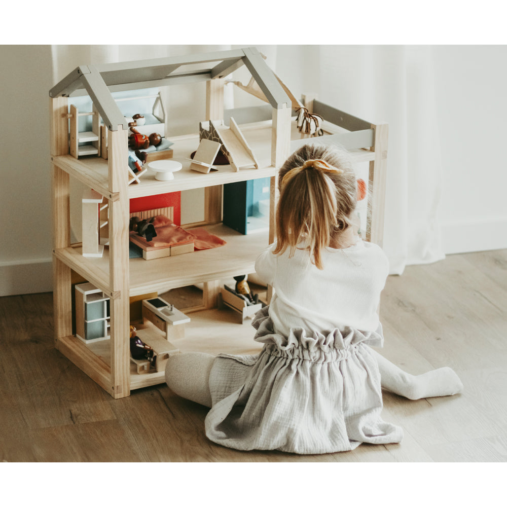 Ein kleines Kind mit Pferdeschwanz spielt mit dem hölzernen Puppenhaus „Puppenhaus Natur mit Möbeln“ auf einem hellen Holzboden in der Nähe eines Fensters.