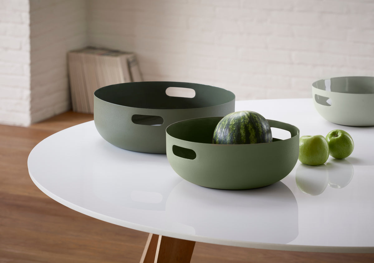 Zwei moderne grüne Schalen auf einem weißen Tisch, eine davon enthält eine Wassermelone, daneben zwei grüne Äpfel.