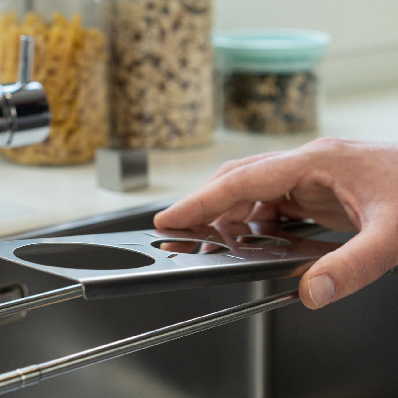 Eine Hand richtet das Metallgestell „Abtropfhilfe Drip.it“ über einer Küchenspüle aus, im Hintergrund sind Gläser mit Nudeln und Snacks zu sehen.