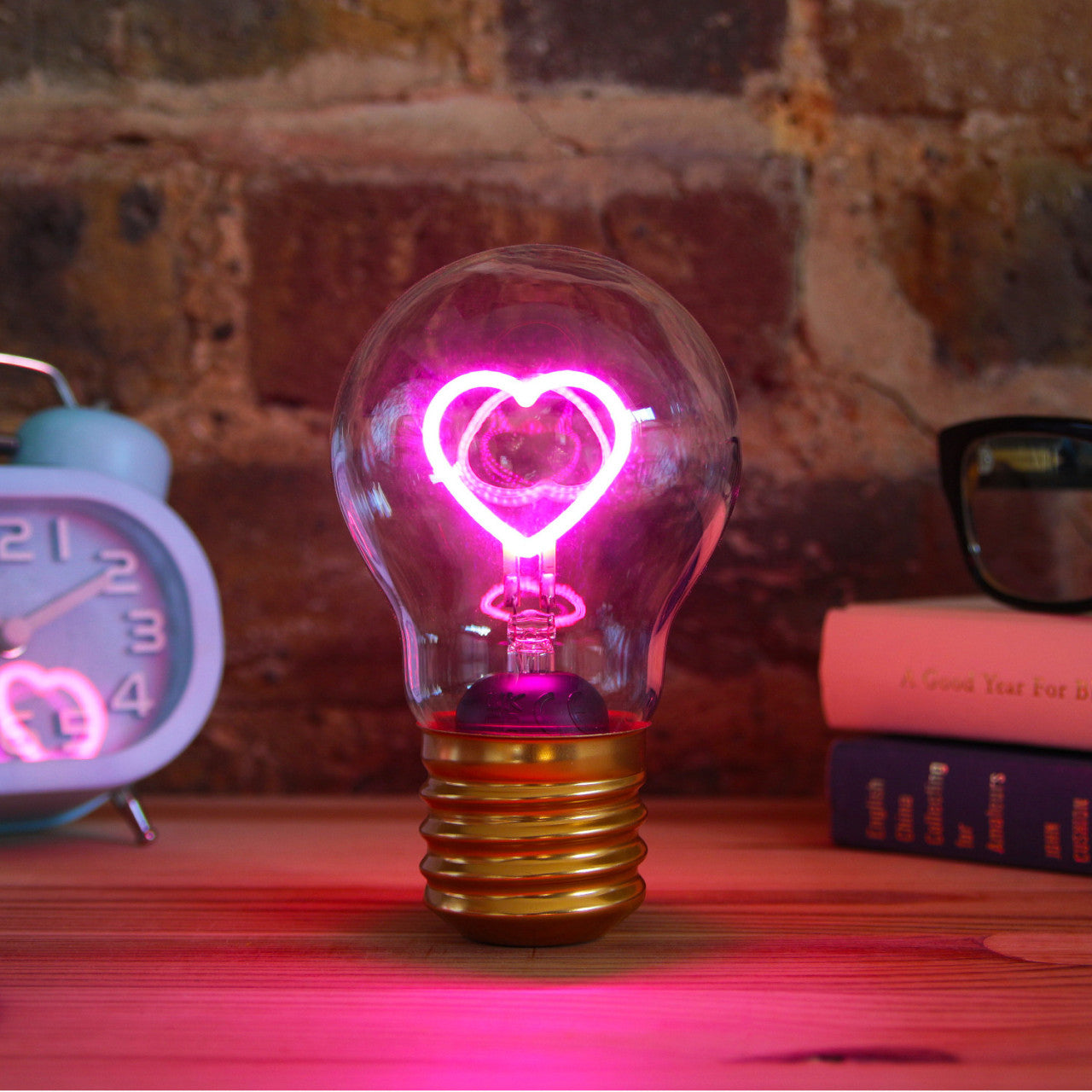 Die Kabellose LED-Glühbirne - Herz leuchtet mit einem rosa, herzförmigen Glühfaden auf einem Holztisch neben einer Uhr und Büchern.