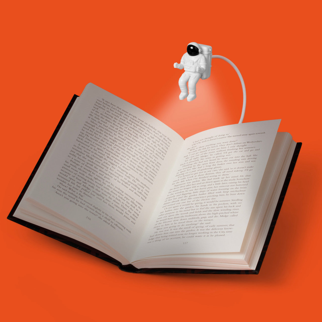 Ein offenes Buch, beleuchtet von der Leselampe Spaceman – 2 LEDs, einer Leselampe in Astronautenform, vor einem orangefarbenen Hintergrund.