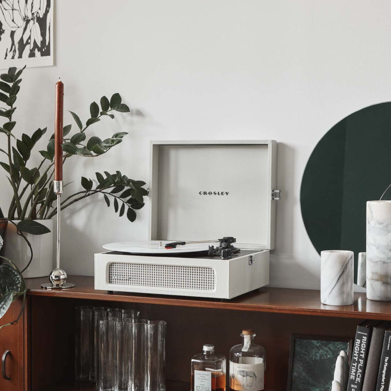 Ein Sandgrau Crosley Voyager Plattenspieler steht auf einem Holzschrank, umgeben von Pflanzen, Kerzen und Glaswaren.