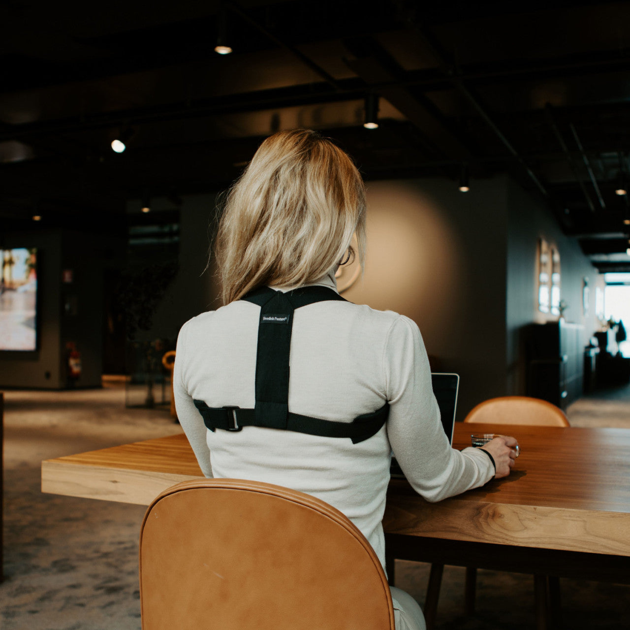 Eine Person mit langen blonden Haaren trägt den Haltungskorrektor Haltungstrainer schwarz M-L, während sie an einem Tisch sitzt und in einem Büro einen Laptop benutzt.