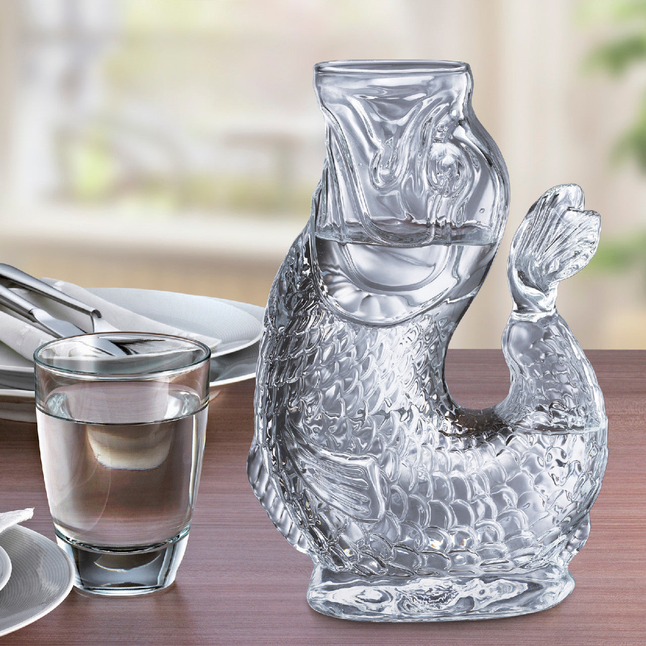 Die Wasserkaraffe NEMO – Transparent, eine fischförmige Wasserkaraffe aus Glas, steht mit einem Glas Wasser auf einem Esstisch.