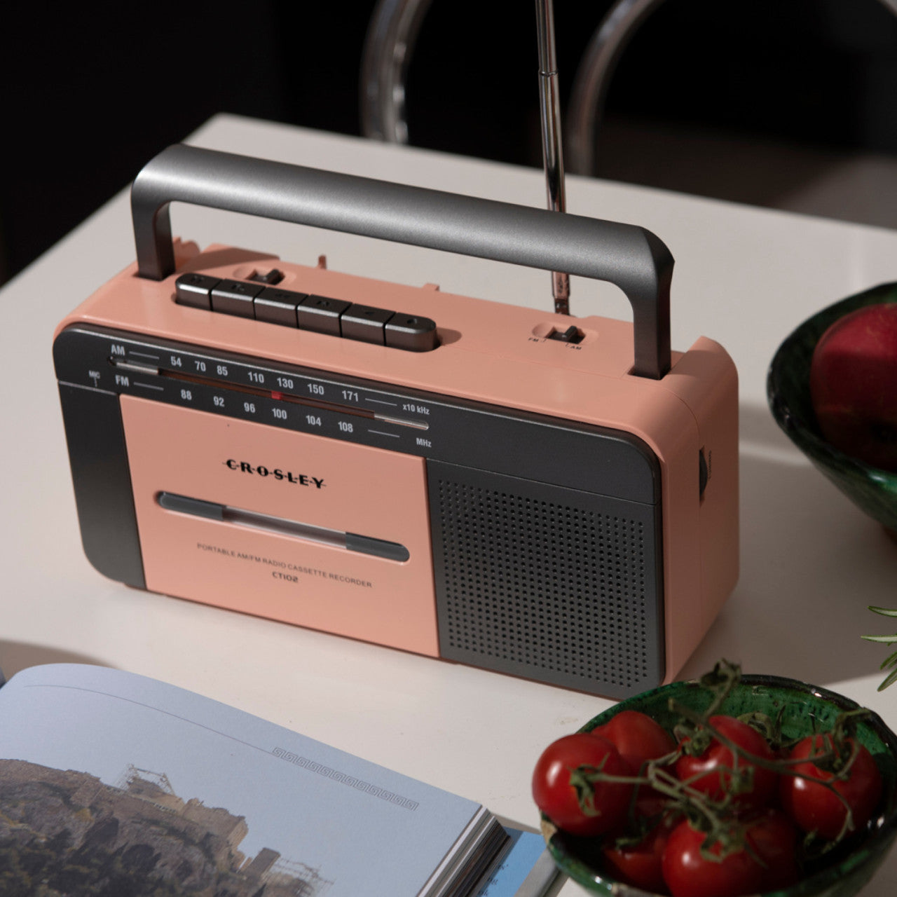 Ein Crosley-Kassettenspieler in Rosa/Grau steht auf einem Tisch neben Tomaten und einer offenen Zeitschrift.