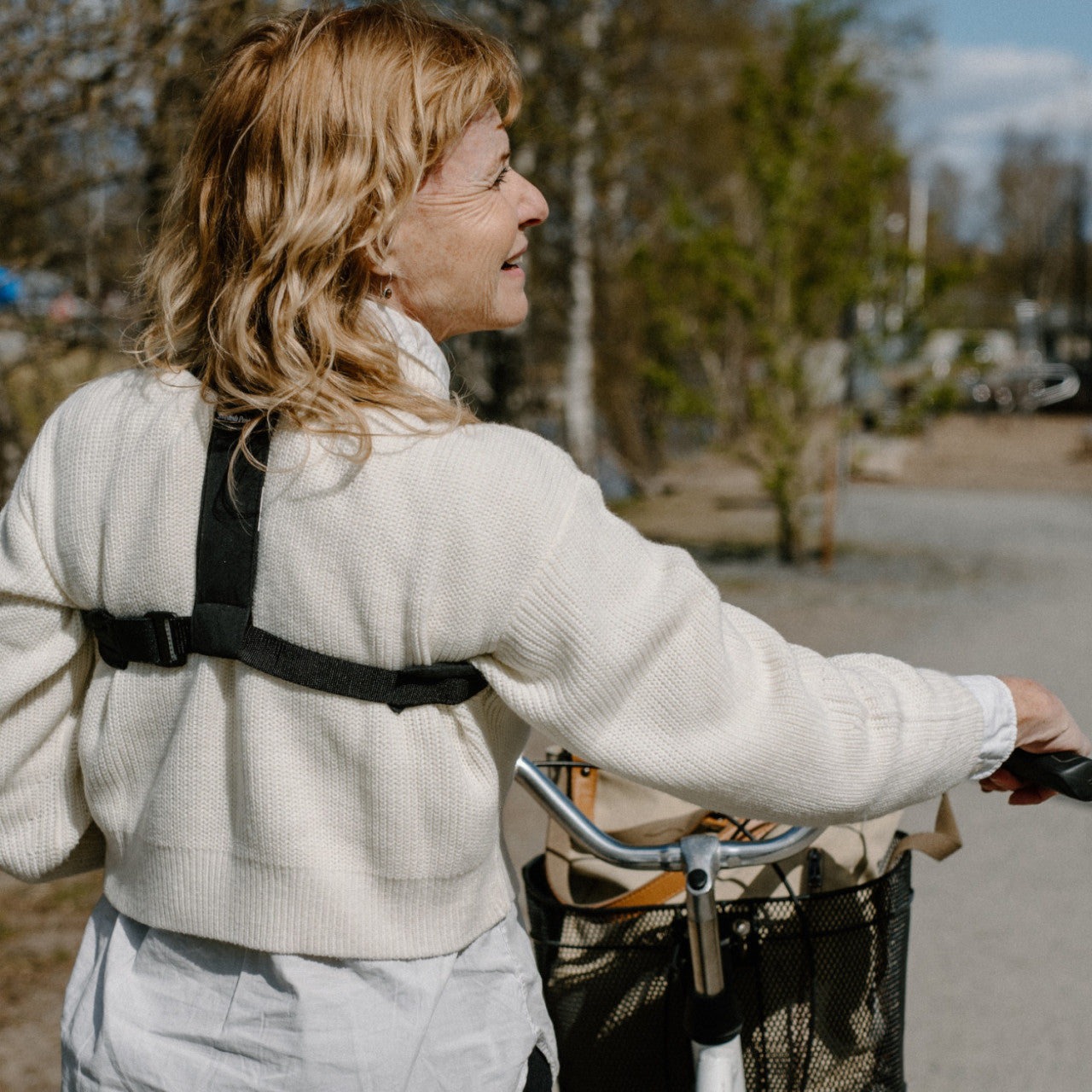 Lächelnde Frau mit blonden Haaren, trägt einen Haltungstrainer schwarz M-L und einen cremefarbenen Pullover, schiebt im Freien ein Fahrrad und schaut zur Seite.