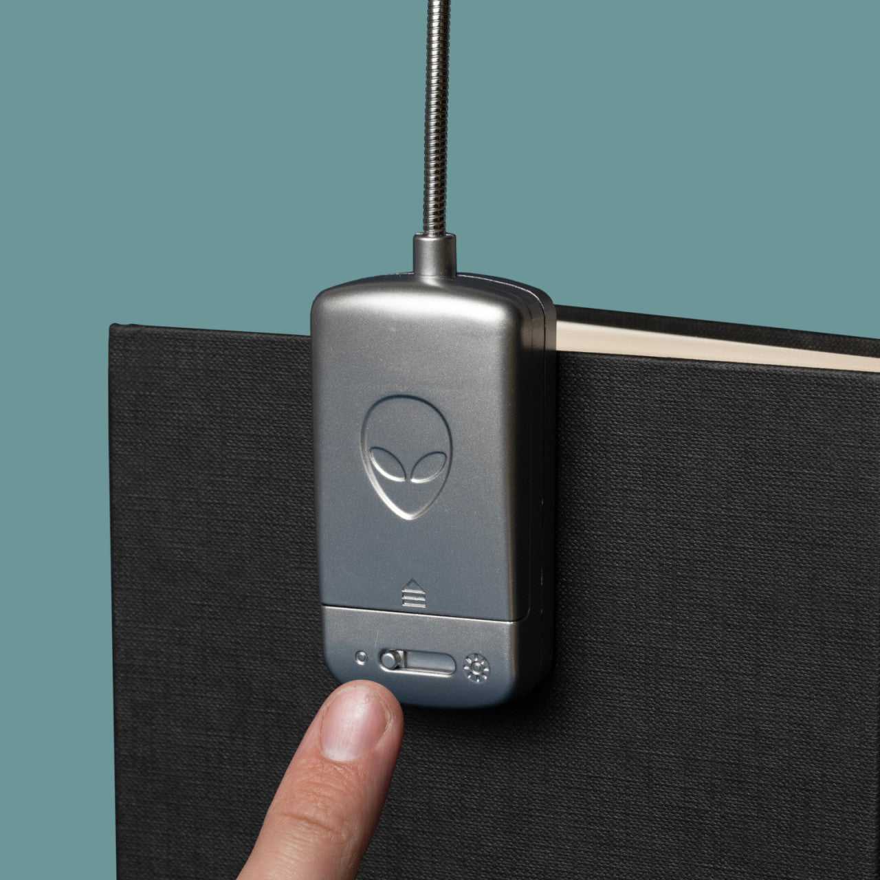 Ein Finger drückt den Knopf der Leselampe UFO – 3 LEDs, ein metallisches Leselicht mit einem Alien-Gesicht-Logo, das an einem schwarzen Buch befestigt ist.