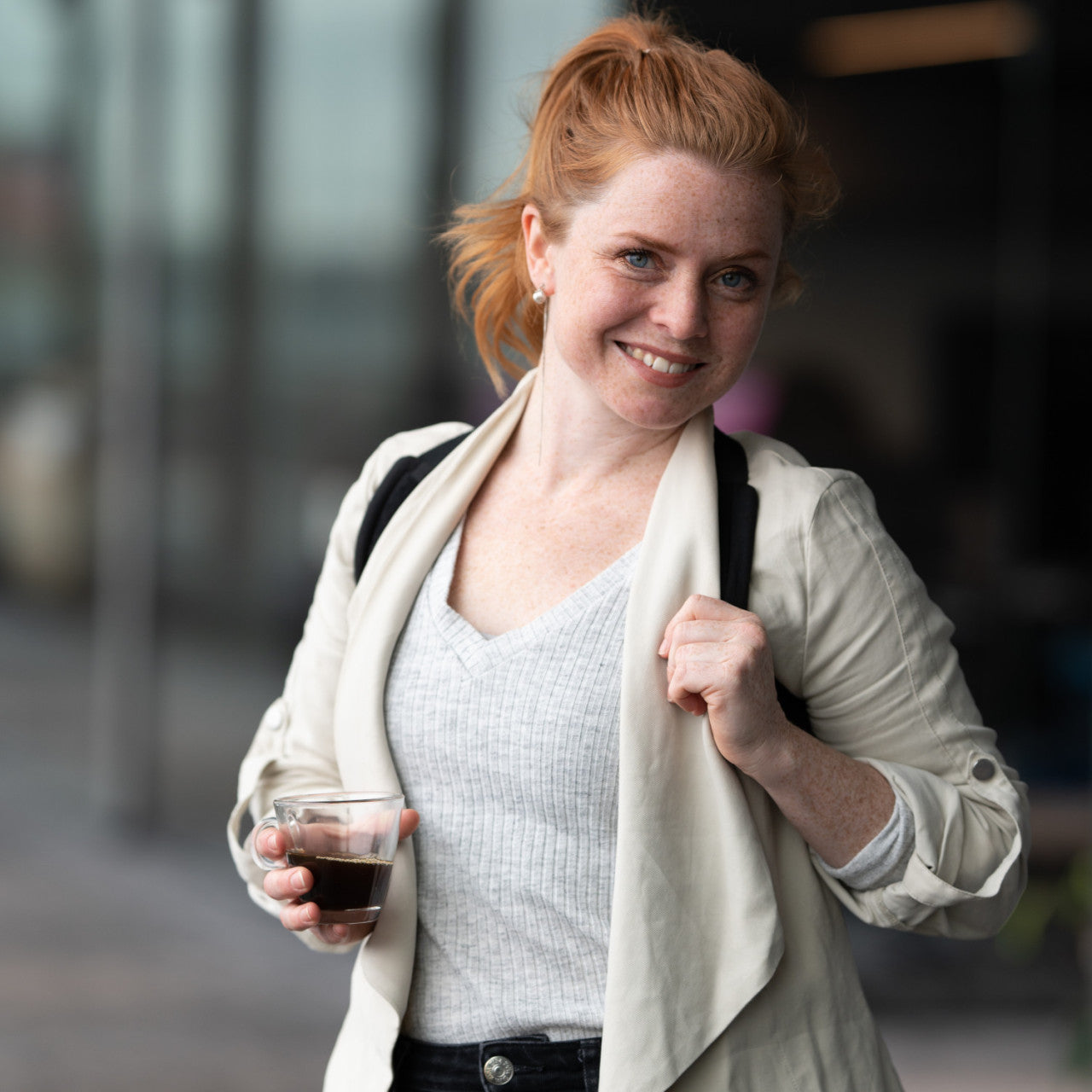 Lächelnde Frau mit roten Haaren, trägt einen Haltungstrainer schwarz M-L unter einem hellen Blazer und hält im Freien ein Getränk.