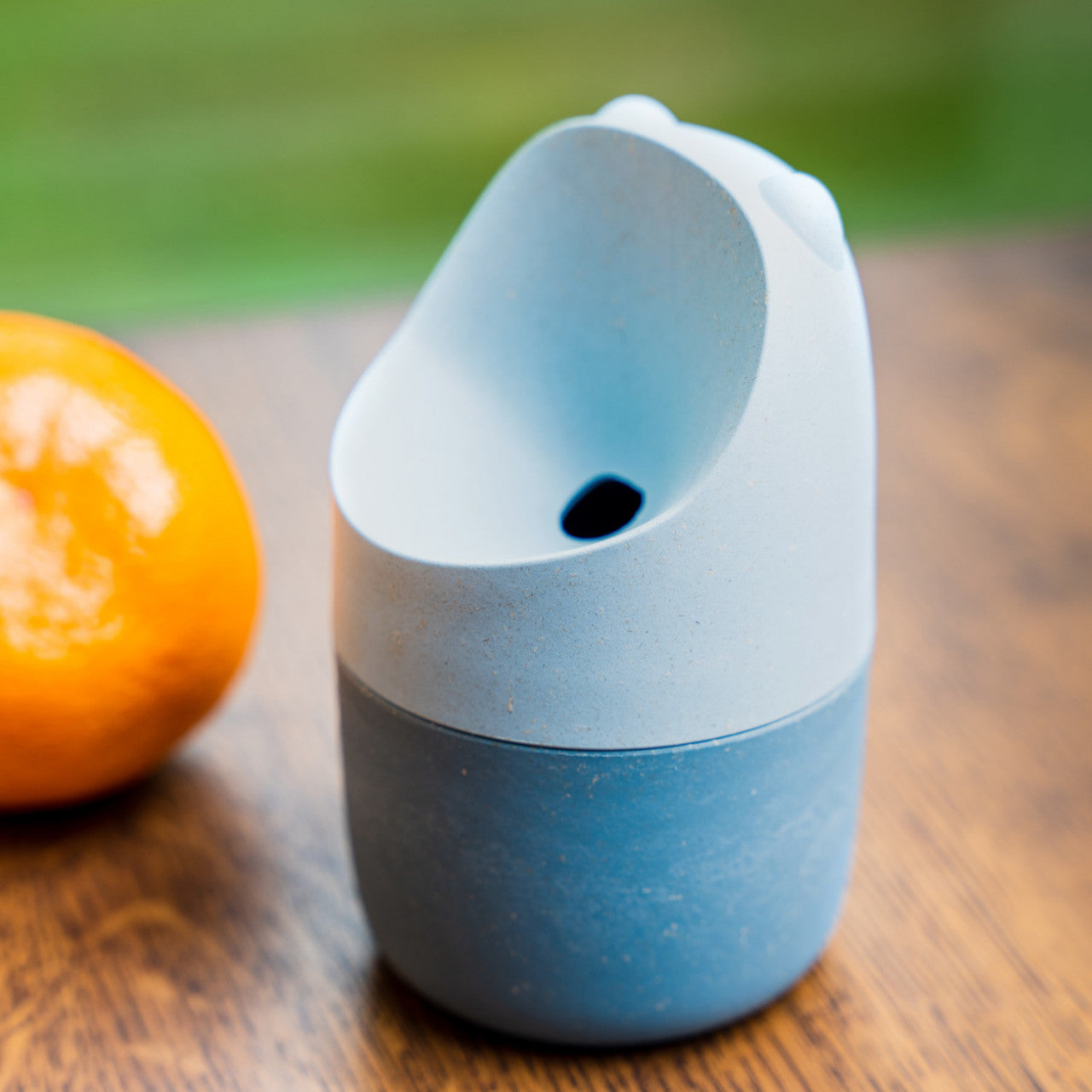 Der Fruchtfliegenfänger KONNA - Blau sitzt auf einer Holzoberfläche neben einer Orange, mit einem unscharfen grünen Hintergrund.