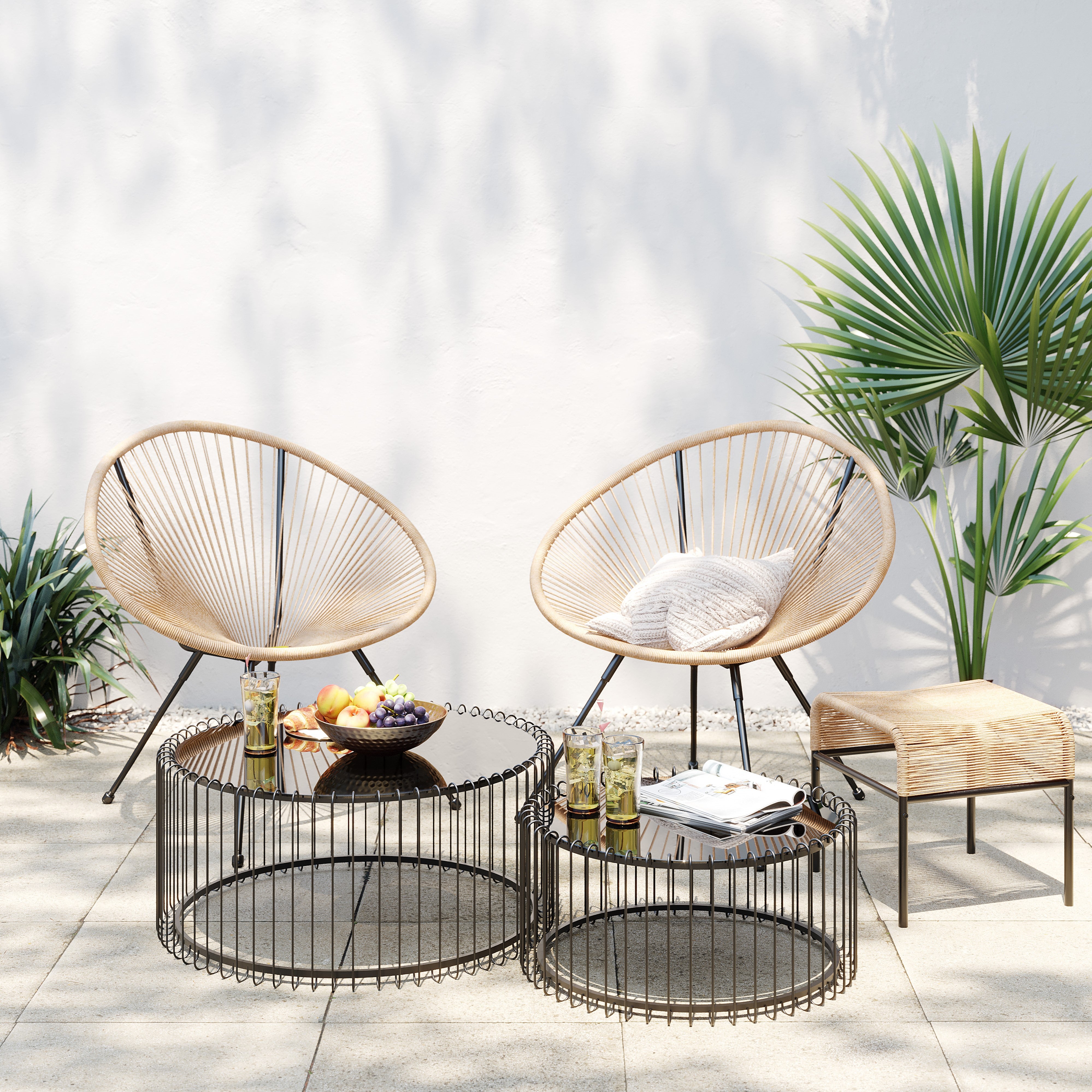 Zwei runde Acapulco Outdoor Stühle mit hellen Kissen, zwei Drahtgestell-Couchtische mit Getränken, einer Obstschale, Zeitschriften und ein Hocker Acapulco schwarz/natur schaffen neben Pflanzen und einer weißen Wand eine einladende Sitzgelegenheit auf einer sonnigen Terrasse.