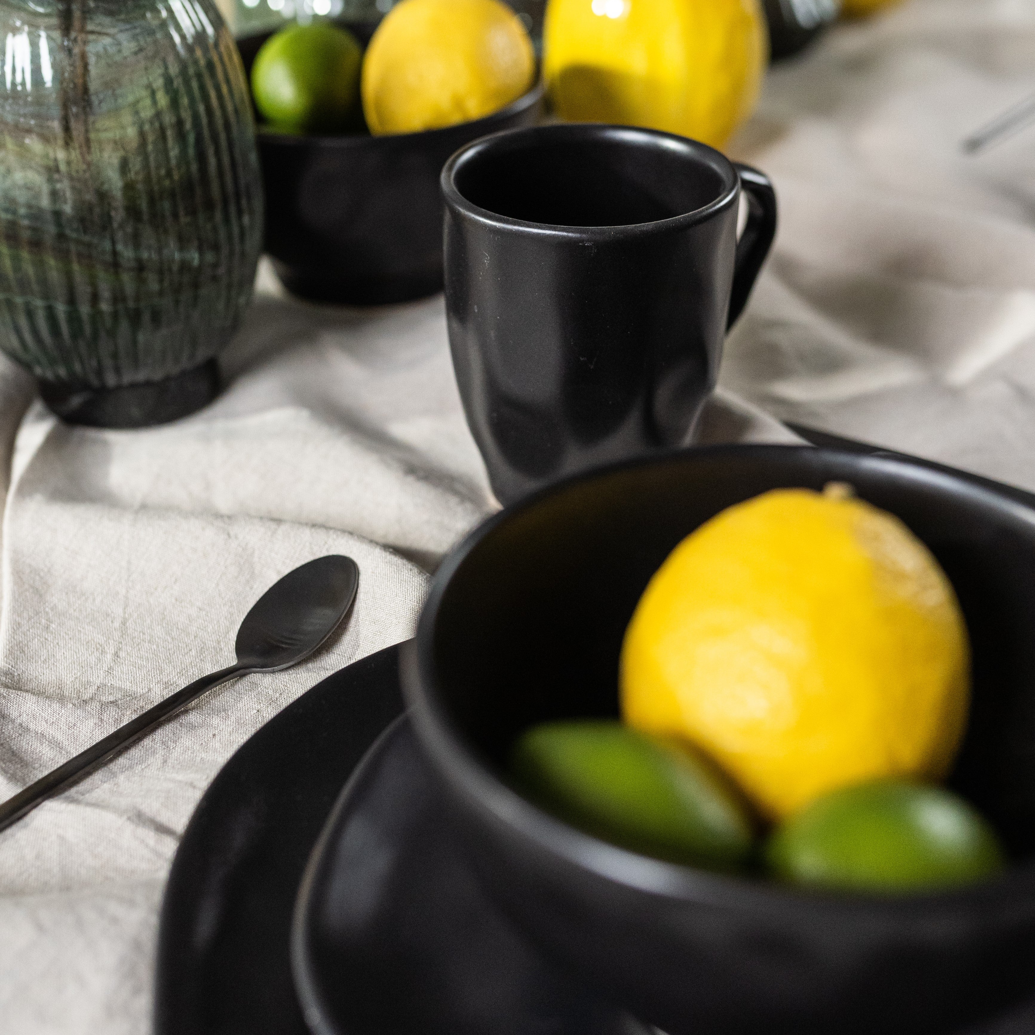 Eine Nahaufnahme eines gedeckten Tisches mit der Tasse Organic Black, schwarzem Steingut-Geschirr, einer Schale mit einer Zitrone und zwei Limetten, einem Löffel und einer grünen Vase auf einer cremefarbenen Tischdecke.