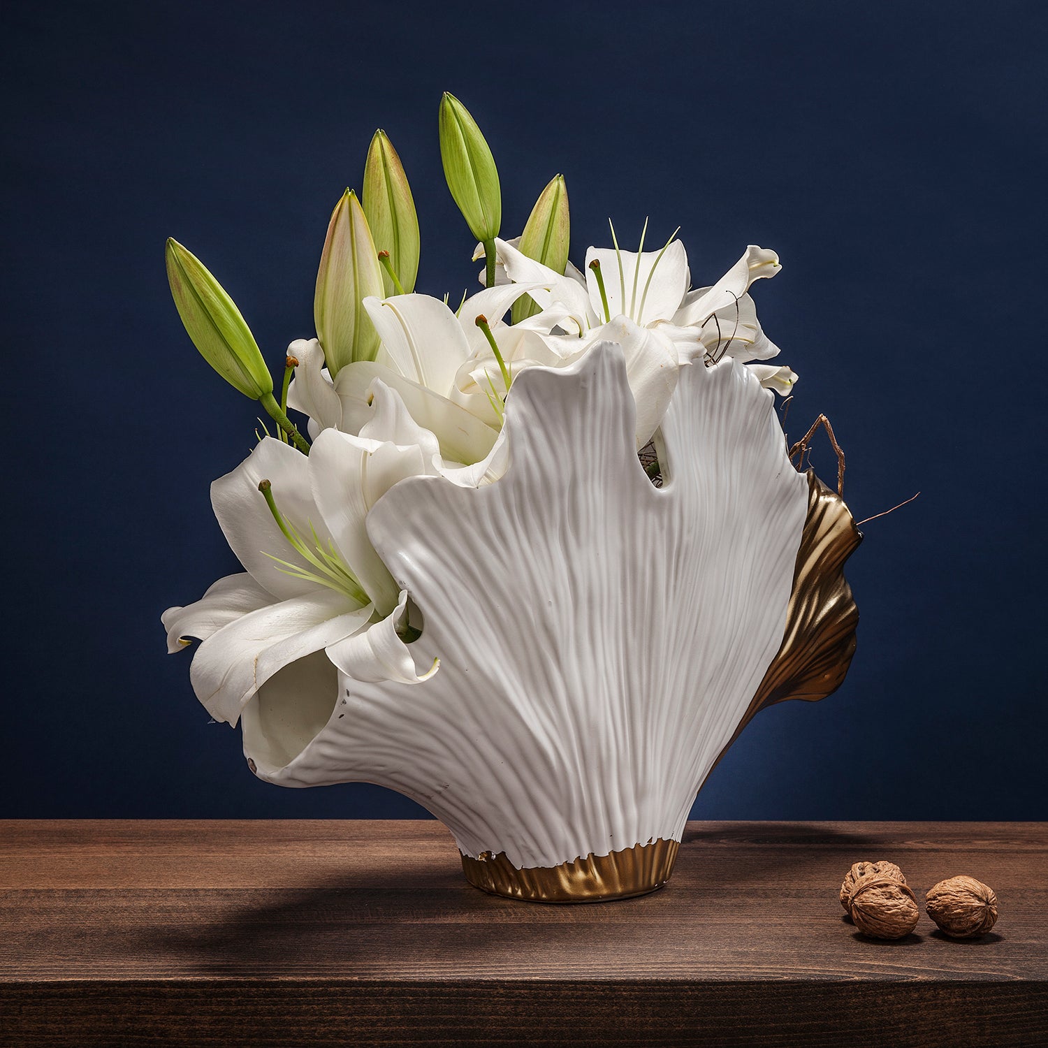 Die Vase Ginkgo Elegance mit ihrem skulpturalen weiß-goldenen Design enthält blühende weiße Lilien und Knospen. Dieses elegante Stück steht auf Holz vor einem dunkelblauen Hintergrund, begleitet von zwei Walnüssen.