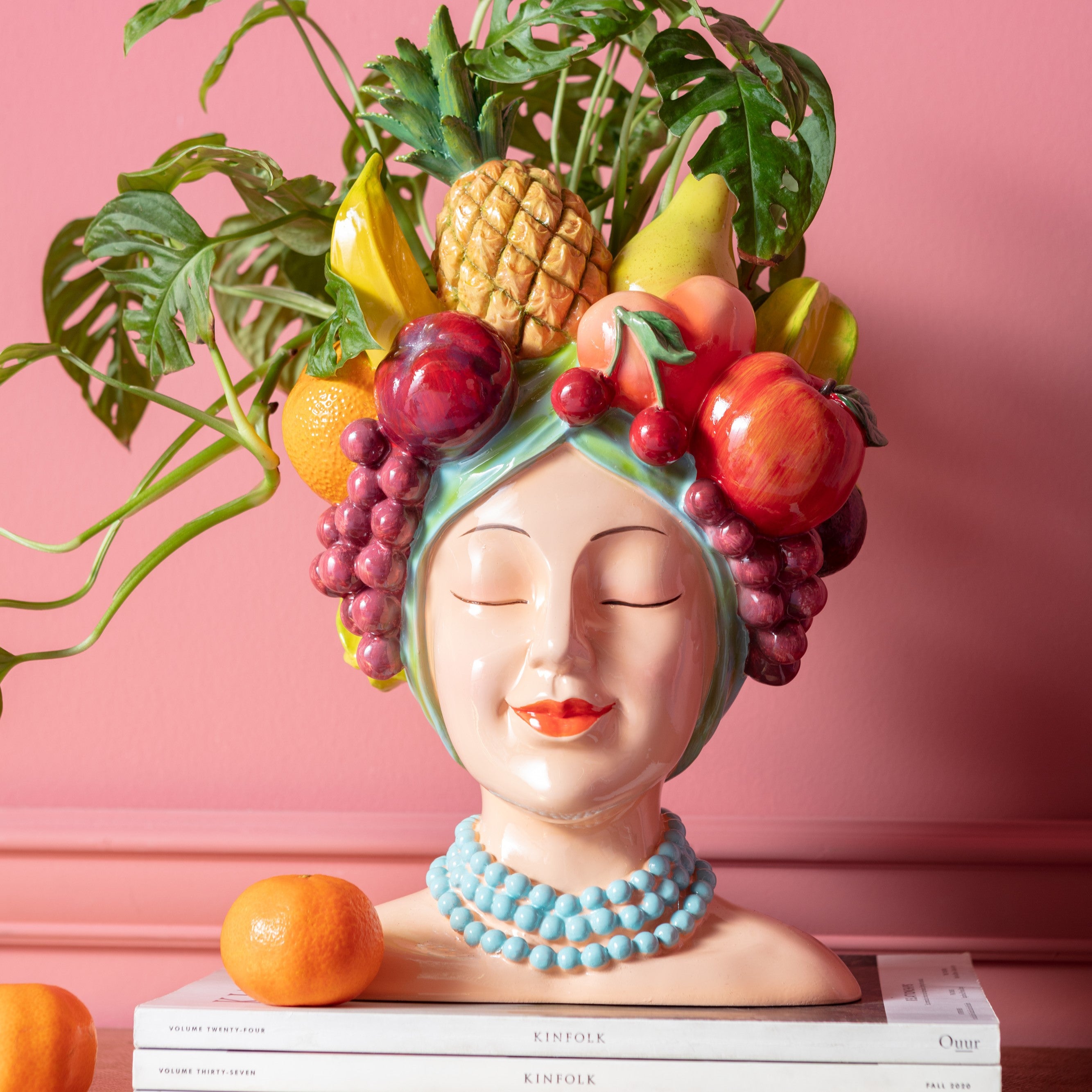 Die Deko Vase Fruity zeigt eine lächelnde Frau mit geschlossenen Augen, einer blauen Perlenkette und einem Fruchtkopfschmuck – eine auffällige Keramikbüste, die sich perfekt als Couchtisch-Deko vor einer rosa Wand mit grünen Blättern, Büchern und einer Orange davor eignet.