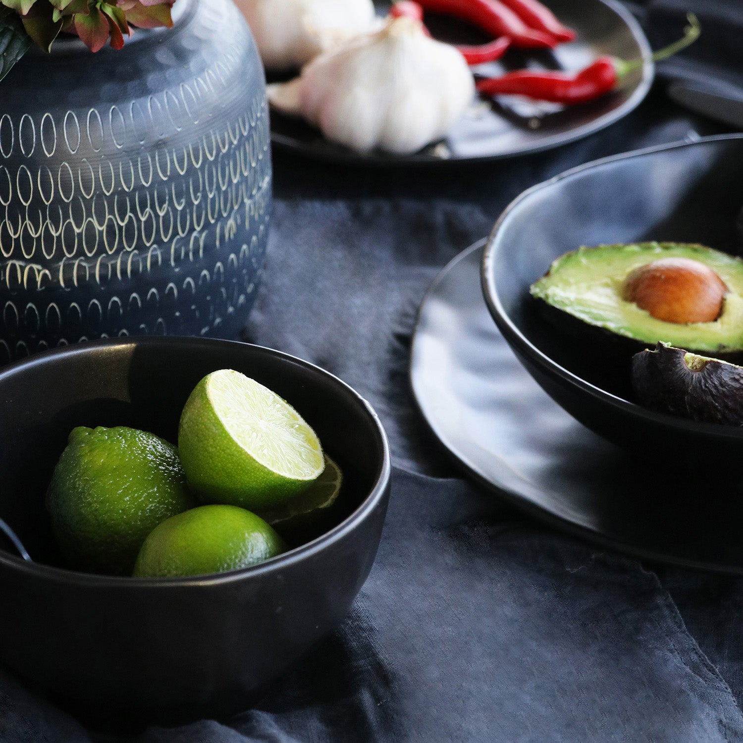 Eine Schale Organic Black (Ø 15 cm) mit Limetten steht auf einer dunklen Tischdecke neben Tellern mit Avocado, Knoblauch und roten Chilischoten. Eine gemusterte Keramikvase ist teilweise sichtbar.